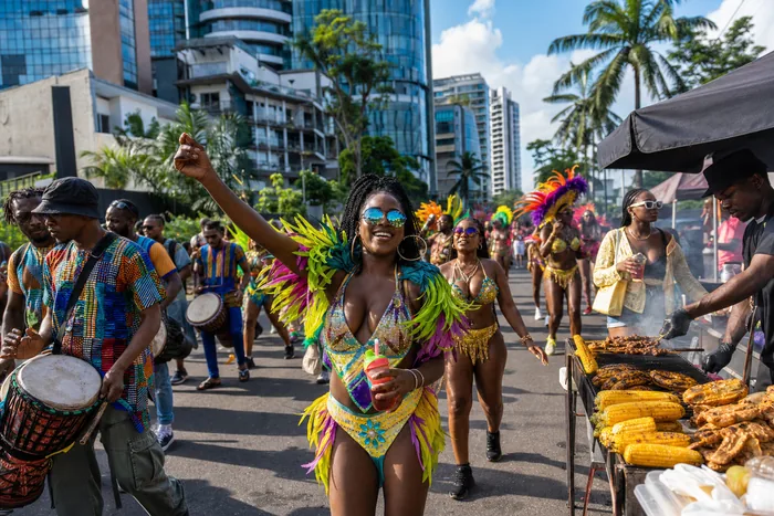 A Lagos street carnival scene in Victoria Island with dancers, carnival bands, food vendors and city buildings in the background.
